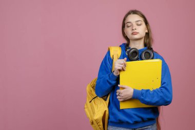Sırt çantalı, kulaklıklı, gözleri kapalı güzel bir kadın. Mavi khudi giyen, elinde klasör tutan güzel bir kız öğrenci. Pembe stüdyo arka planında izole edilmiş..