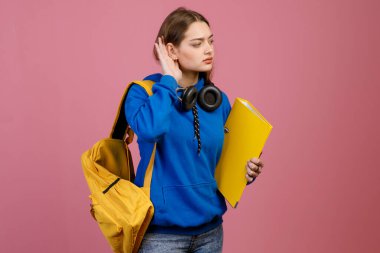 Ayakta duran, kulağına dokunan, dinleyen güzel esmer kızın ön görüntüsü. Kulaklıklı, sarı sırt çantası ve klasörü olan güzel bir öğrenci. Pembe stüdyo arka planında izole edilmiş..