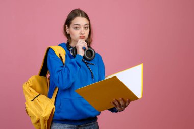 Önde duran güzel kız öğrenci, elinde klasör, kitap, düşünme. Güzel esmer kız kameraya bakıyor, çenesine dokunuyor. Pembe stüdyo arka planında izole edilmiş..