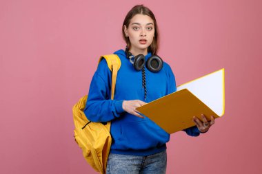 Güzel liseli kızın ağzı açık kameraya baktığı ön manzara. Sarı açık klasör ve sırt çantası tutan esmer kadın, günlük kıyafetler giyiyor. Gençlik kavramı.