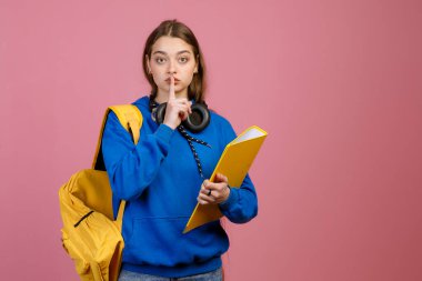 Önde duran esmer, güzel kız öğrenci, sessizliğini koruyor. Kulaklıklı, sarı sırt çantalı, klasörlü, kameraya bakan güzel bir kadın. Pembe arka planda izole edilmiş.