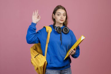 Önde duran esmer kız, elini kaldırıyor, iyi görünüyor. Sarı sırt çantası ve klasör tutan güzel kız öğrenci kameraya bakıyor. Pembe arka planda izole edilmiş.