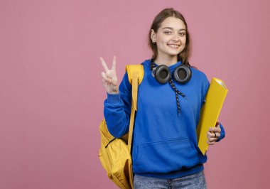 Esmer kızın ayakta duruşu, gülümsemesi, barışı göstermesi, selamlaması. Sarı sırt çantalı ve dosyasıyla kameraya bakan güzel kız, mutlu. Eğitim ve gençlik kavramı.