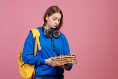 Front view of surprised schoolgirl holding bunch of old books. Pretty brunette female standing, looking down, wearing fashionable clothes. Isolated on pink studio background. clipart