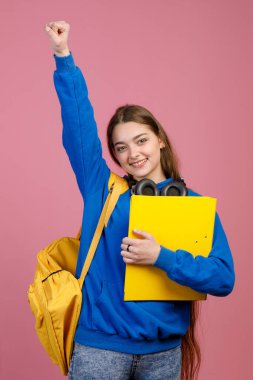 Kulaklıklı, ayakta duran, elini kaldıran mutlu kız öğrencinin ön görüntüsü. Sarı klasör ve sırt çantasıyla gülümseyen güzel bir bayan. Gençlik ve üniversite yaşam tarzı kavramı.