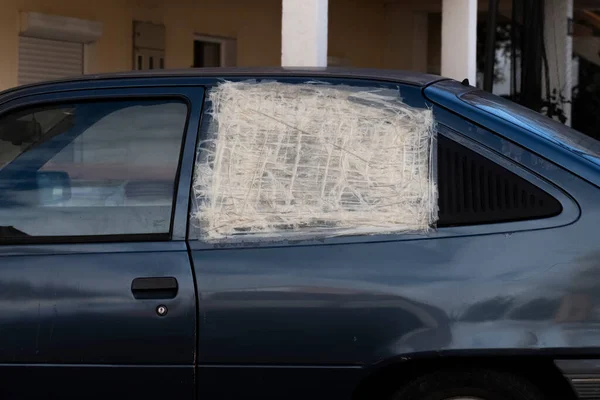 Yolcu kapısında kırık cam olan beyaz bir araba beyaz filme sarılmış ve beyaz boya bandıyla mühürlenmiş. Yeşil yapraklı eski bir hurda araba..