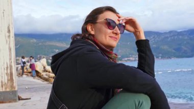 Güneş gözlüklü bir kızın deniz kenarındaki plajda oturup manzaraya hayran olduğu yakın plan. Denizde tatil. Dünyayı dolaş. Kız denizde günbatımının tadını çıkarıyor..