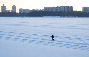 Şehirdeki donmuş bir nehrin geniş bir yatağında karda kayak yapan bir kayakçının silueti. Bir kış günü gün batımında evlerin arasında yürü..