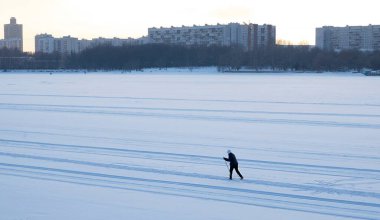 Şehirdeki donmuş bir nehrin geniş bir yatağında karda kayak yapan bir kayakçı figürü. Sıcak giysili bir kadın kış günü gün batımında evlerin arasında kayak yapıyor..