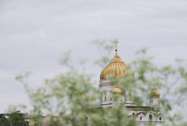 The outline of the large golden-domed dome of the Orthodox Christian Church of Christ the Savior through the trees and green foliage on a summer day. Lots of white light clouds in the sky.