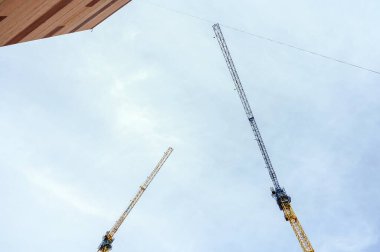 Parlak güneşli bir gökyüzüne karşı mavi ve sarı renkli iki uzun kule inşaat vinci. Yüksek katlı bir tuğla evin yanında..