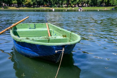 Güneşli yaz havasında parktaki suyun yüzeyine ahşap bir tekne boyadı. Tekne bir iple iskeleye bağlandı..