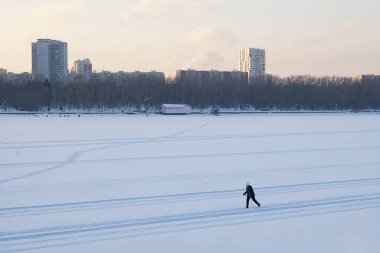 Moskova, Rusya. 26.12.2021. Bir kayakçı şehir merkezindeki donmuş bir nehrin geniş bir yatağında karda kayak yapıyor. Sıcak giysili bir kadın kış günü gün batımında evlerin arasında kayak yapıyor..