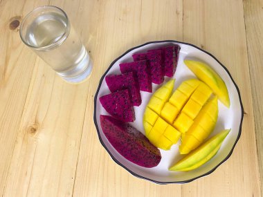 Ahşap zemin üzerinde bir bardak suyla bir tabak sağlıklı mango ve ejderha meyvesi dilimleri. Üst görünüm.