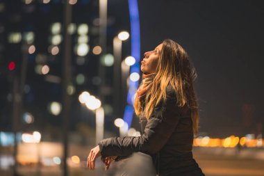 Yalnız bir kadın poz veriyor ve düşünceli. Hiçbir yere bakmıyor. Arka planı bulanık bir şehir mumuyla aydınlatılmış.