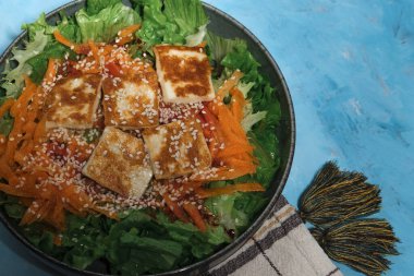 Havuçlu yeşil salata ve kızarmış halloumi peyniri, mutfak tekstil peçetesi, mavi desenli arka planda servis edilir.