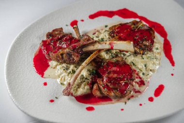 Kırmızı kiraz soslu ızgara kuzu pirzola. Yulaf lapası ve mevsimlik otlarla kaplı. Beyaz tabak ve arka plan beziyle servis ediliyor.