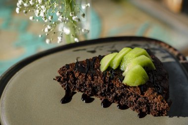 Çikolatalı brownie taze kivi dilimleriyle servis edilir, güneşli bir kafede modern seramik kil tabakta.