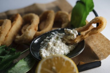 Kızarmış kalamar halkaları, sosla servis edilen altın kahverengi kabuklu yiyecekler, yeşiller ve limon, yemek kağıdıyla şekillendirilmiş.