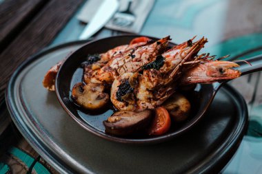 Izgara jumbo karides güveci ya da güveçte domatesli, tereyağlı sarımsak soslu ve mantarlı dünya renginde kahverengi tabaklar servis ediliyor.