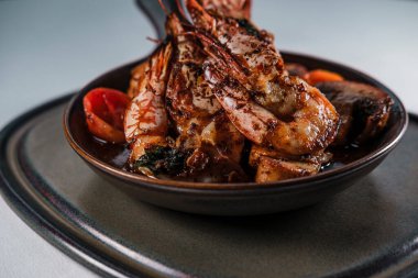 Izgara jumbo karides güveci ya da güveçte domatesli, tereyağlı sarımsak soslu ve mantarlı dünya renginde kahverengi tabaklar servis ediliyor.