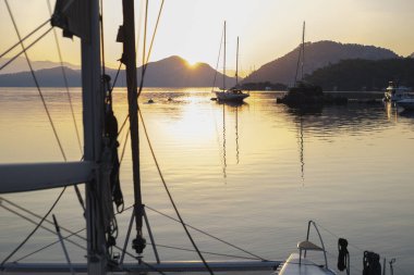 Sabahın sakin manzarası güverteden sakin gün doğumu demirleme yerinde, yükselen güneş, dağlar, deniz ışık yansımaları, alacakaranlık renkleri, yelkenli botlar, dingin liman