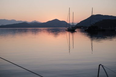 Sabahın sakin manzarası güverteden sakin gün doğumu demirleme yerinde, yükselen güneş, dağlar, deniz ışık yansımaları, alacakaranlık renkleri, yelkenli botlar, dingin liman