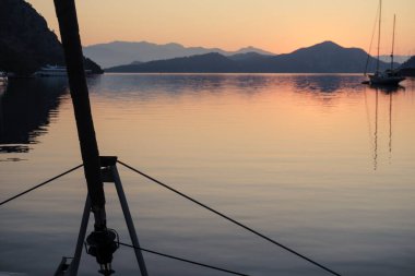 Sabahın sakin manzarası güverteden sakin gün doğumu demirleme yerinde, yükselen güneş, dağlar, deniz ışık yansımaları, alacakaranlık renkleri, yelkenli botlar, dingin liman