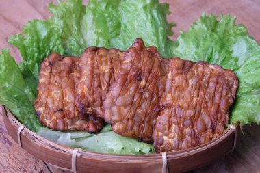 Tempe goreng, kızarmış tempeh. Tempeh ya da tempe mayalanmış soya fasulyesinden yapılan geleneksel Endonezya yemeğidir. Tahta masadaki bambu dokuma kabında marulun üzerinde. Üçgen şekilli tempeh. Süper yemek..