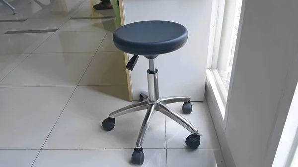 A rolling stool with black wheels by the window. A cushioned seat. Stylish bar stool with wheels.
