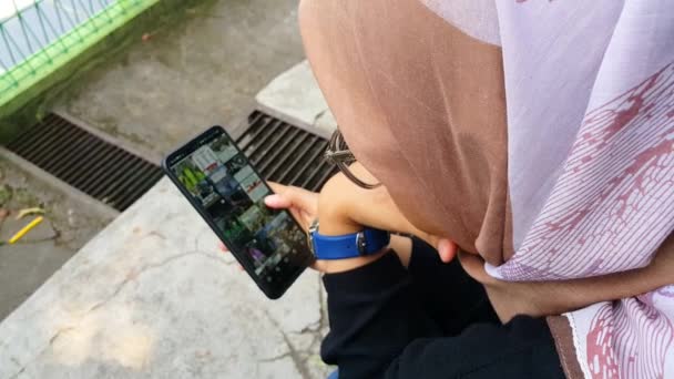一个年轻的亚洲穆斯林妇女 戴着头巾 眼镜和手表 在户外用智能手机 一个年轻的穆斯林妇女在智能手机屏幕上滚动 — 图库视频影像