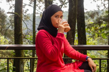 Asyalı Müslüman bir kadın balkonda oturmuş buzlu kahve içiyor. Arka planda doğa ya da ağaçlar var.