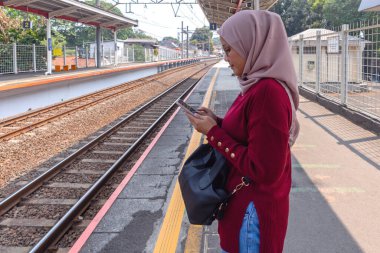 Asyalı Müslüman bir kadın trenin gelmesini beklerken cep telefonunu kullanıyor..