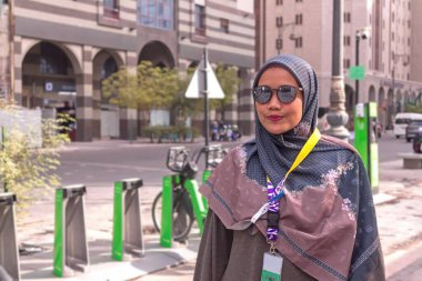 Umrah yolcu kartı ve güneş gözlüğü takan Asyalı Müslüman bir kadın, Suudi Arabistan 'ın Medine şehrinde bir şehir caddesinde poz veriyor.