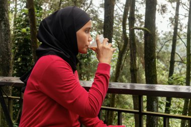 Müslüman bir kadın balkonda oturup içki içiyor. Arka planda doğa ya da ağaçlar var.