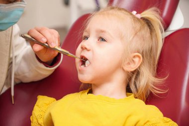 Kız dişçi koltuğunda oturuyor, doktor çocuğun ağız boşluğunda çeşitli manipülasyonlar yapıyor..