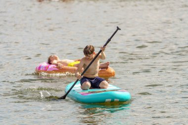 Bir çocuk denizde şişme bir tahtanın üzerinde yüzüyor ve kardeşini şişme bir yüzüğe bindiriyor..
