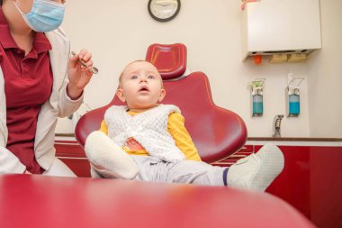 Küçük bir çocuk diş muayenesi koltuğunda oturmuş diş hekiminin aletlerini tutan yardımcısına tuhaf bir şekilde bakıyor. Klinik parlak ve donanımlı, hastalara dostane bir ortam sağlıyor..
