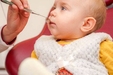 Küçük bir çocuk dişçi koltuğunda oturmuş elinde bir enstrüman tutan diş hekimine bakıyor. Klinik aydınlık ve renkli, dostane bir ortam yaratıyor..
