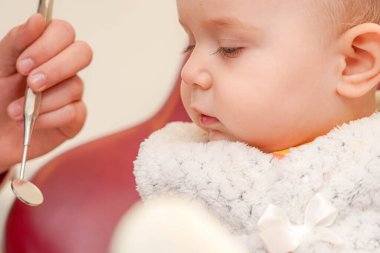 Küçük bir çocuk sakin sakin otururken, dişçi dişlerini, rahat bir klinik ortamında diş aynasıyla incelemeye hazırlanıyor ve pediatrik tedaviye nazikçe yaklaşımını gösteriyor..