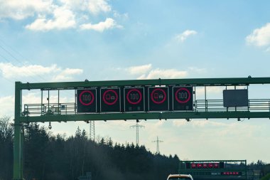 Dijital trafik işaretleri otoyolda hız limitinin 100 olduğunu gösteriyor. İşaretler mavi gökyüzüne karşı açıkça görülebiliyor. Dağılmış bulutlar sürücülere önemli bilgiler sağlıyor..