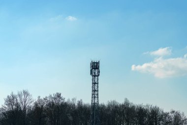 Hücresel bir kule berrak mavi bir gökyüzüne doğru yükseliyor. Baharın başlarını gösteren seyrek ağaçlardan oluşan bir zemin var. Ortam sakin ve doğadaki telekomünikasyon altyapısını gösteriyor..
