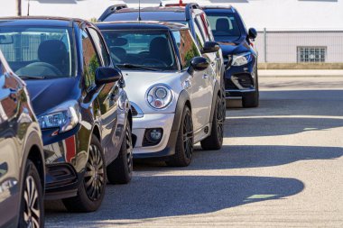 Sakin bir muhitte güneşli bir günde araçlar sıraya dizilir. Arabaların parlak tasarımları, temiz asfalt yolu ve yukarıdaki parlak mavi gökyüzü ile tezat oluşturarak, huzurlu bir ortam yaratıyor..