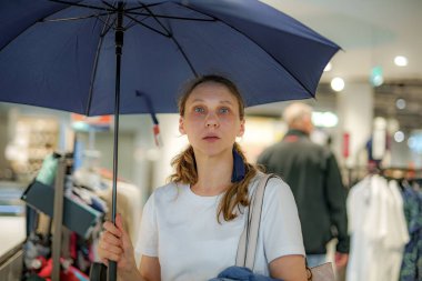 Bir kadın elinde mavi bir şemsiyeyle perakende mağazasında duruyor, diğer müşterilerin arasında şaşkın şaşkın bakıyor. O koridorda yürürken parlak ışıklar arka planı aydınlatır..