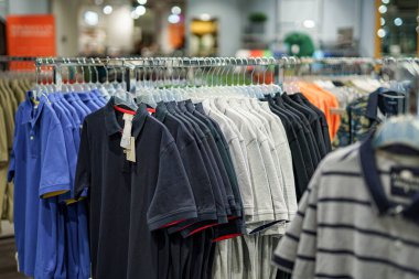 Renkli polo tişörtlerin sıraları bir perakende mağazasında asılı, çeşitli stiller ve renkler sergileniyor. Müşteriler koridorları canlı bir atmosferde gezerler..
