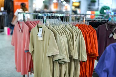 Sıralar halinde düzenlenmiş polo tişörtler çeşitli renklerde sergilenmekte olan bir mağazanın vitrininde asılı duruyor. Müşteriler seçime canlı bir atmosferde bakıyorlar..