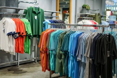 Farklı renklerde polo gömlekleri bir perakende mağazasında raflarda düzgünce asılı duruyor. Müşteriler canlı seçenekleri gözden geçirir, parlak öğleden sonra ışığının tadını çıkarırlar..