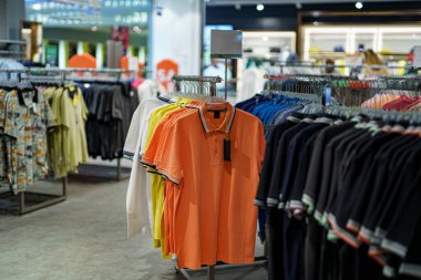 Çeşitli renklerde polo tişörtlerden oluşan canlı bir çeşitlilik perakende mağazasında raflarda asılı duruyor. Müşteriler sezonluk satışlardan yararlanarak seçmelere göz atıyorlar..