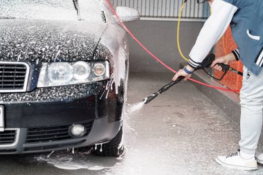 Bir kişi siyah bir aracı basınç makinesiyle temizliyor ve kalın bir köpük tabakası oluşturuyor. Aydınlık bir günde arka planda ekipmanlar olan bir oto yıkama istasyonu..