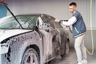 Genç bir adam yan aynaya odaklanmış köpükle bir arabayı temizliyor. Araba yıkama ortamında parlak bir ışıklandırma ve aracın üzerinde köpüklü bir dış görünüş vardır..